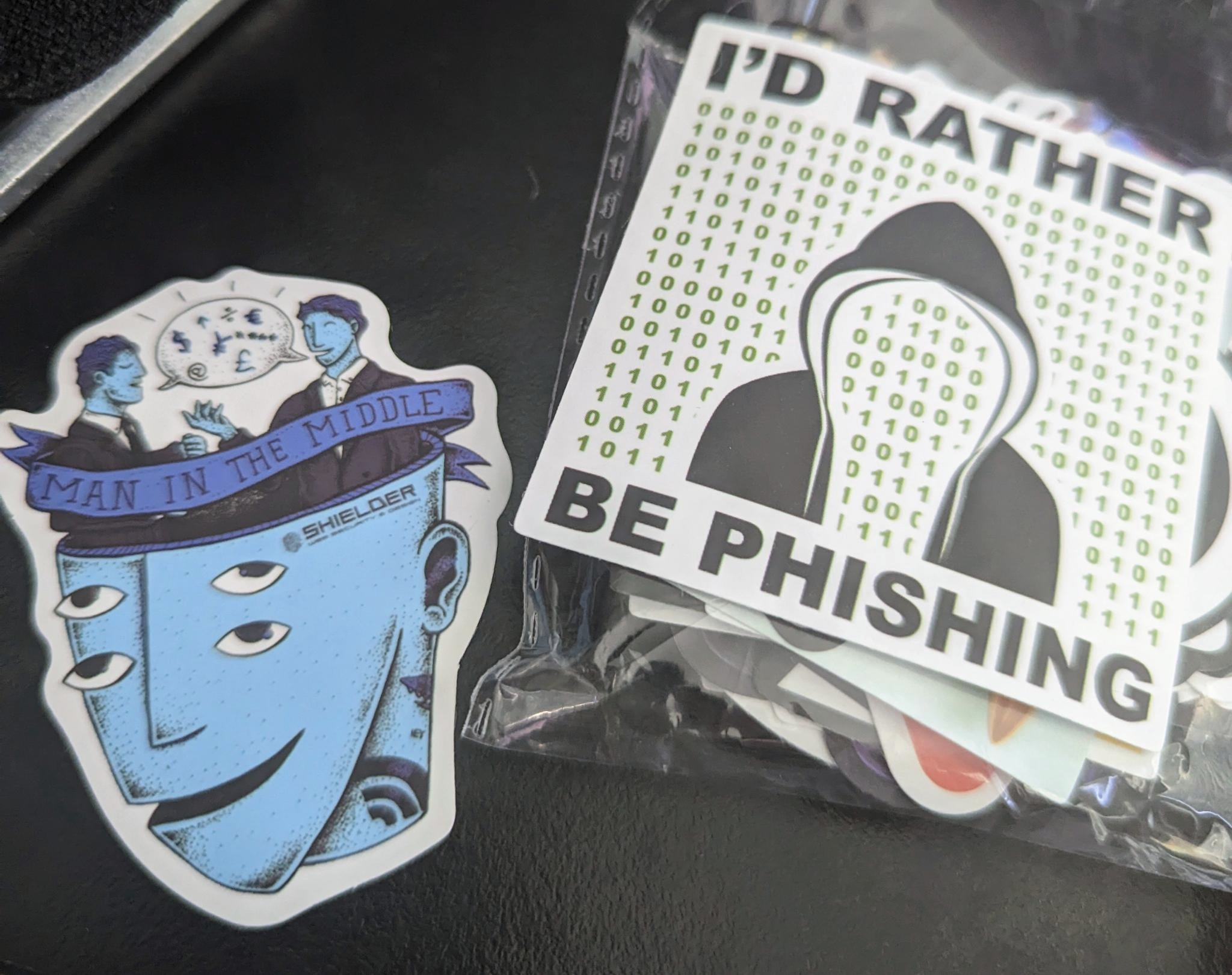 Hacker Stickers. One showing a man in the middle attack depicted with two people conversing inside of a larger third persons head. The second sticker has binary and a hoodie outline with the words &ldquo;I&rsquo;d rather be phishing&rdquo;